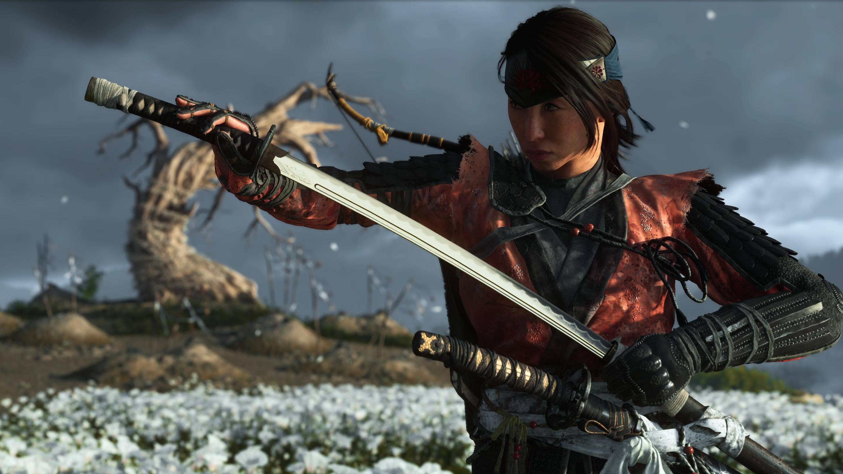 A screenshot of main protagonist Atsu in the action role-playing game Ghost of Yotei, drawing her sword from her scabbard while standing in a field of white flowers and a withering tree in the background.