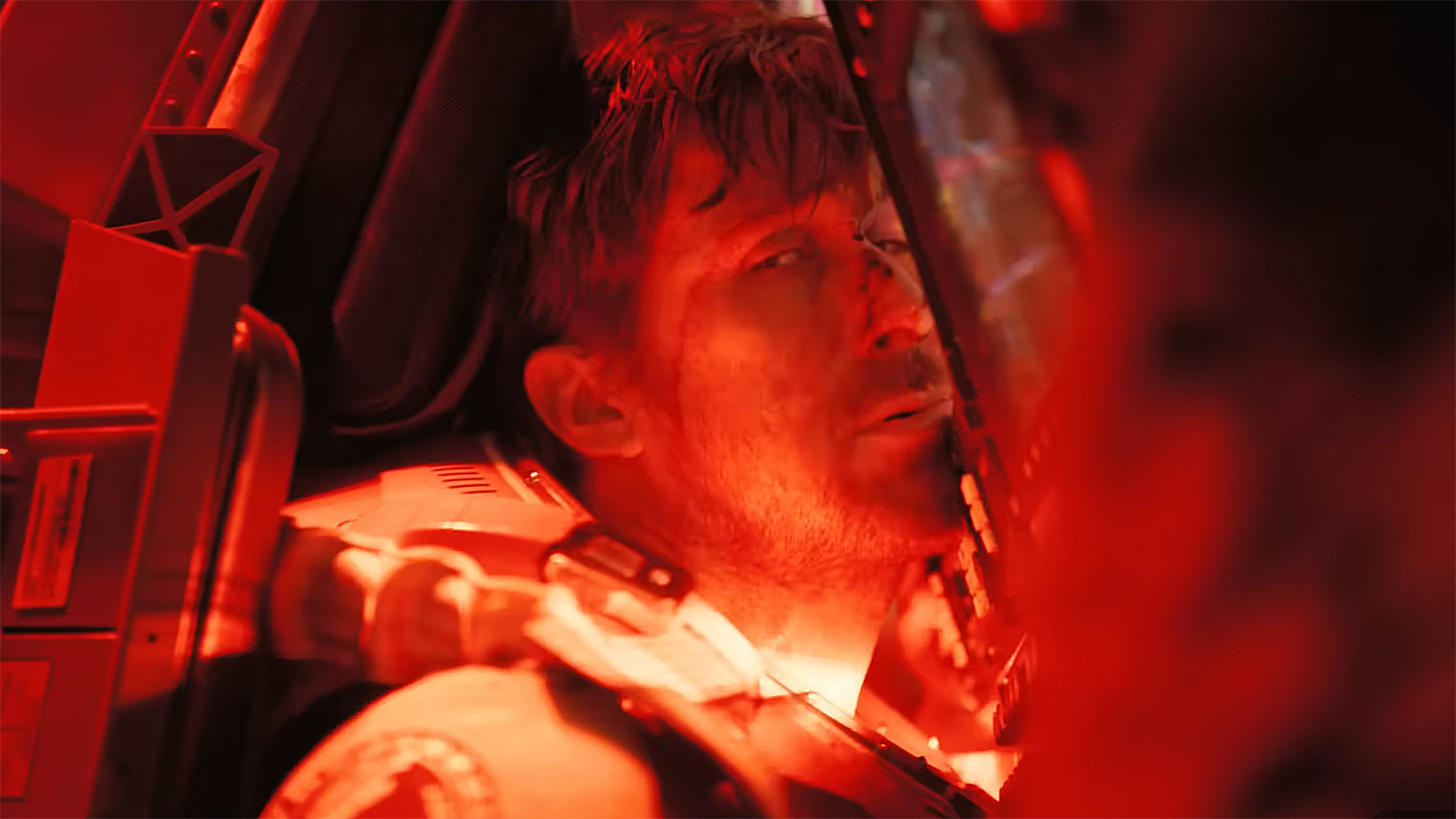 An injured Ryland Grace sitting in a seat pressed up against a spaceship console lit in red light in Project Hail Mary