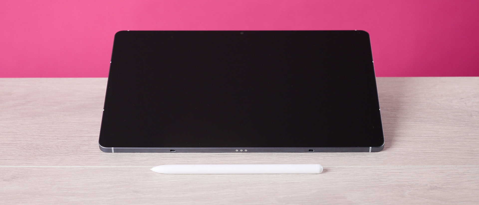 The Samsung Galaxy Tab S10 FE sat on a table behind the S Pen and in front of a pink background.